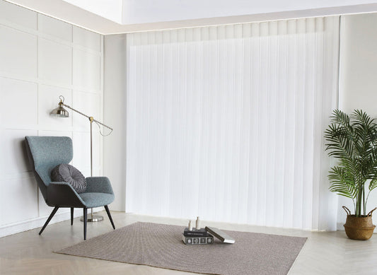 Luxx Shades Plain in white on a floor-to-ceiling window beside an armchair and floor lamp