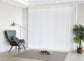 Luxx Shades Plain in white on a floor-to-ceiling window beside an armchair and floor lamp